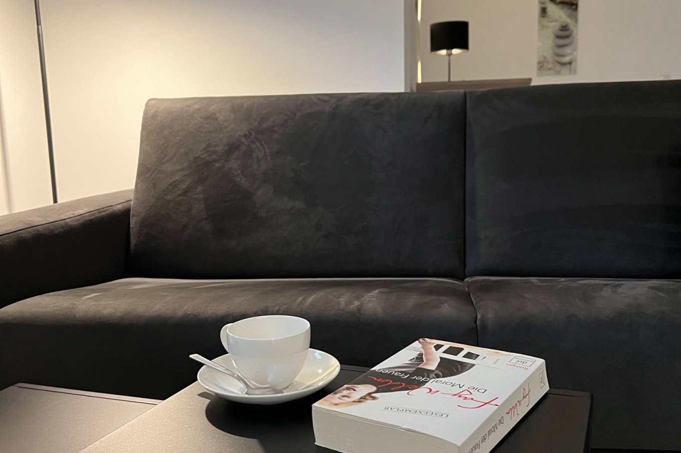 Apartment 1, Wohnzimmer Schwarzes Sofa mit Buch und Tasse auf einem Tisch. Warmes, minimalistisches Ambiente.